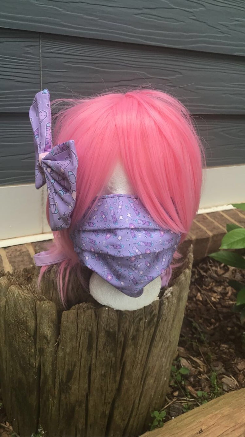 May include: A pink wig with a purple and white bow with a syringe and bandage pattern. The wig is wearing a purple face mask with a syringe and bandage pattern.