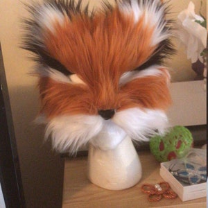 May include: A fluffy orange and white fox head mask with black eyes and a black nose. The mask is on a white mannequin head.