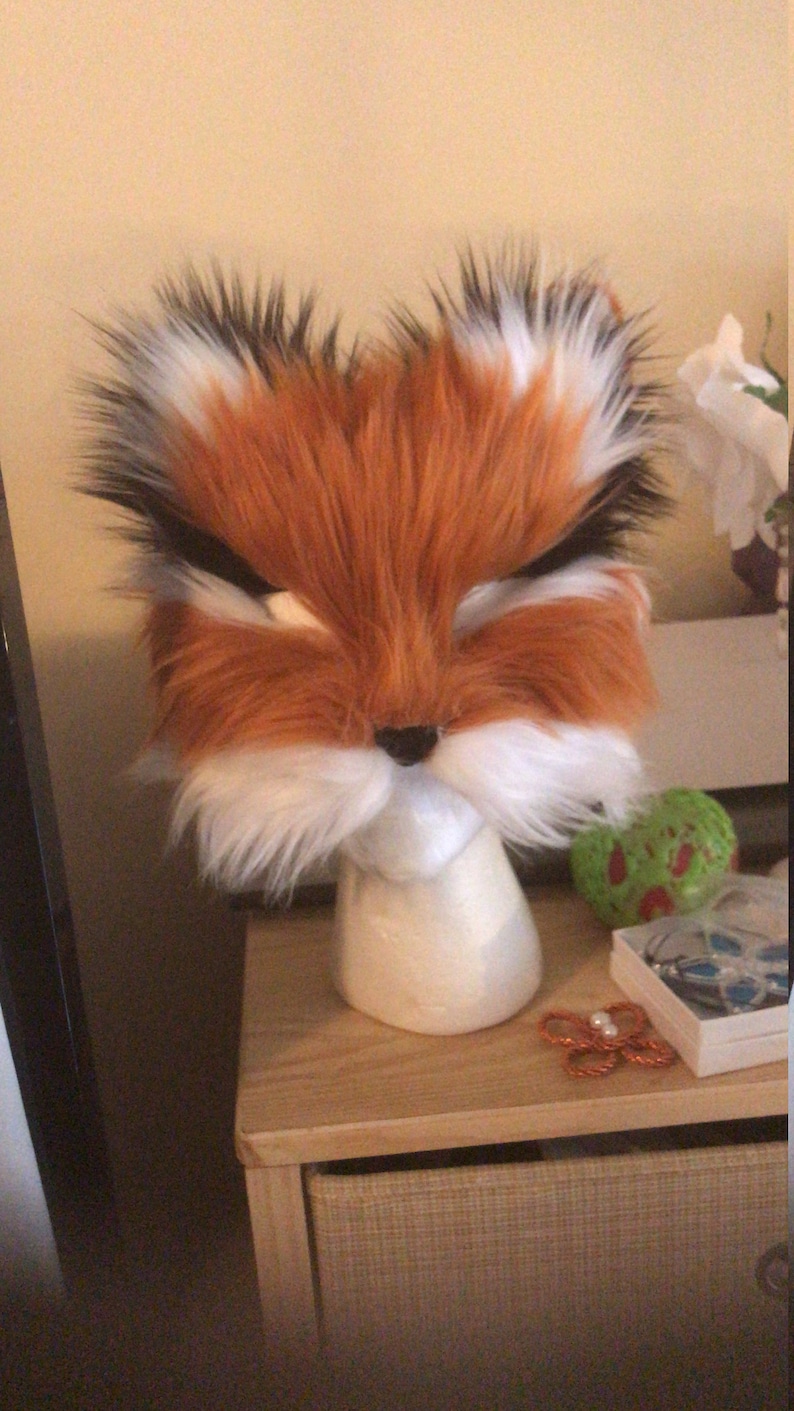 May include: A fluffy orange and white fox head mask with black eyes and a black nose. The mask is on a white mannequin head.