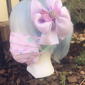 May include: A light blue wig with a large purple satin bow with a jeweled butterfly accent. The wig is styled with a pink face mask with a pattern of eyes, hearts, and bats.