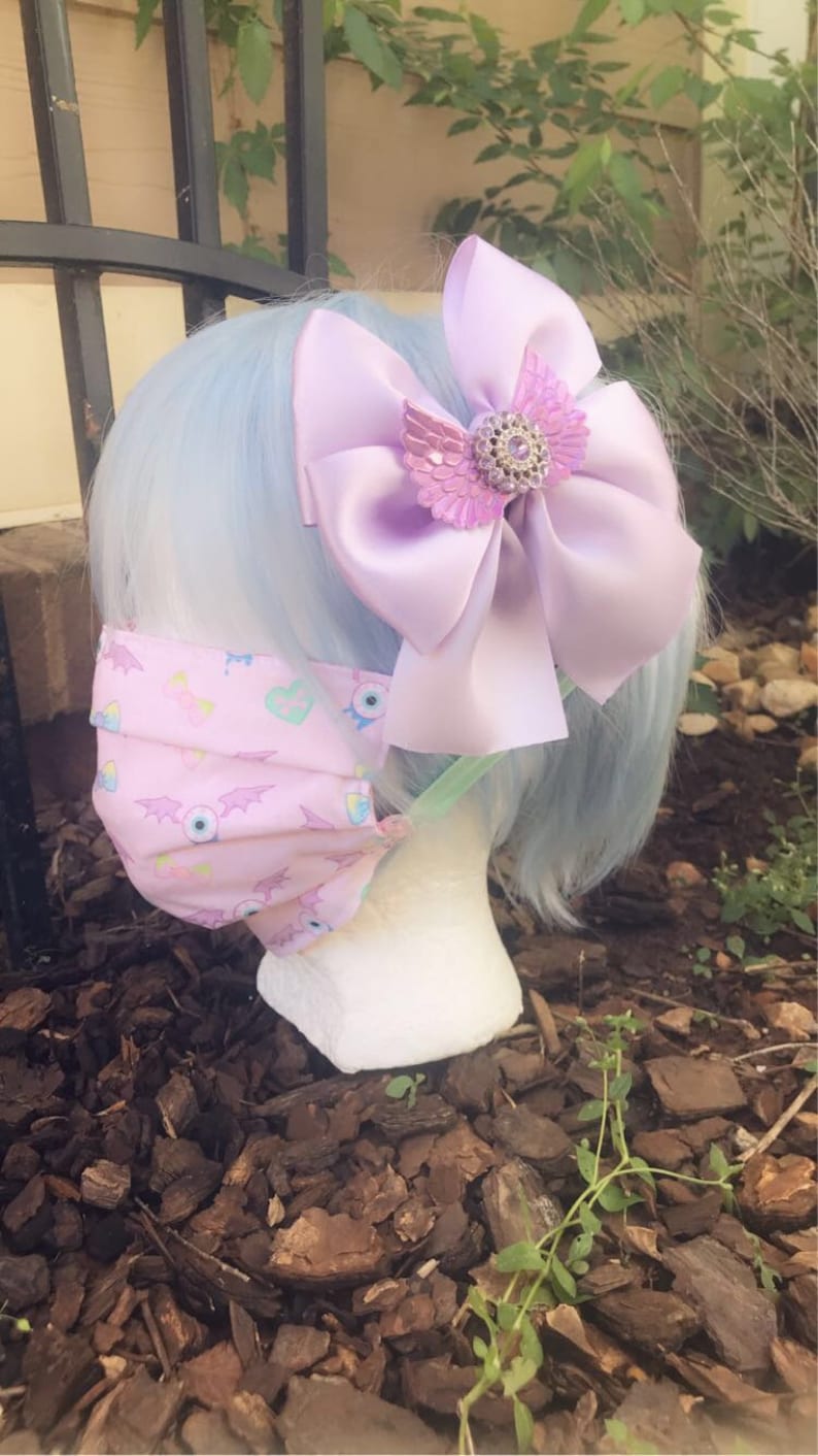 May include: A light blue wig with a large purple satin bow with a jeweled butterfly accent. The wig is styled with a pink face mask with a pattern of eyes, hearts, and bats.