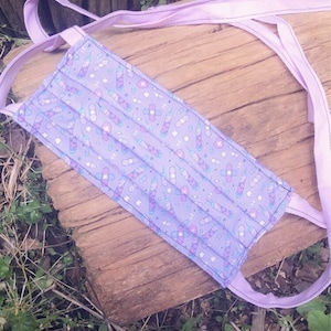 May include: A purple face mask with a white and blue print of pills and bandages. The mask has two long purple ties.