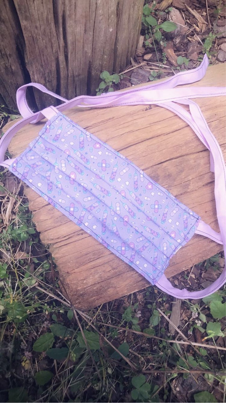 May include: A purple face mask with a white and blue print of pills and bandages. The mask has two long purple ties.