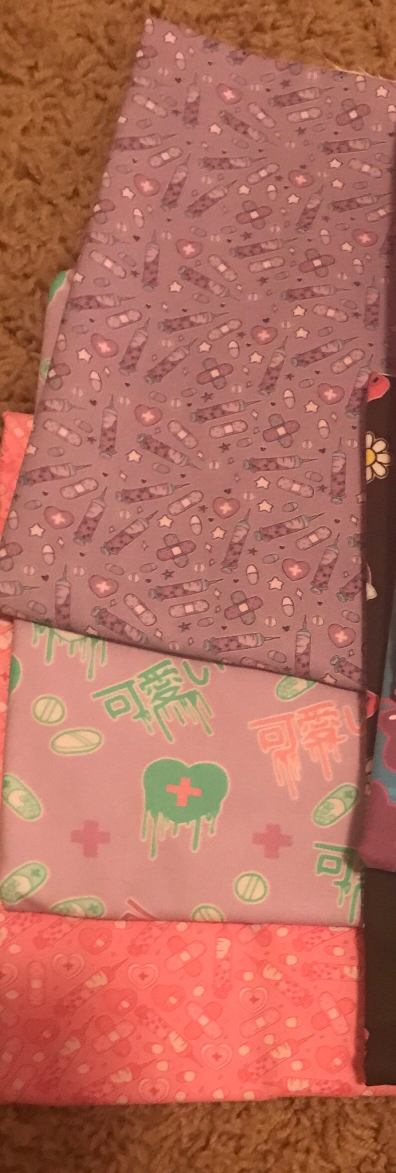 May include: A stack of three fabric squares with different patterns. The top square is purple with a repeating pattern of white syringes, bandages, and pills. The middle square is purple with a repeating pattern of green hearts, white pills, and white plus signs. The bottom square is pink with a repeating pattern of white hearts, bandages, and pills.