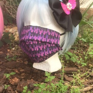 May include: A blue wig with a black bow and a pink butterfly hair clip. The wig is on a mannequin head wearing a purple and black face mask with a dripping pattern.