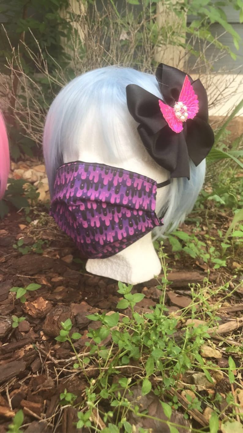 May include: A blue wig with a black bow and a pink butterfly hair clip. The wig is on a mannequin head wearing a purple and black face mask with a dripping pattern.