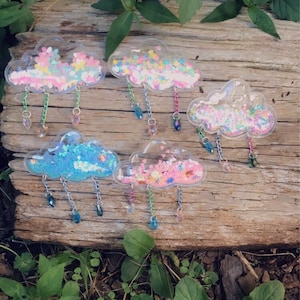 Op de afbeelding: Vijf transparante, wolkvormige broches van plastic met verschillende kleuren glitter erin. Elke broche heeft een ketting met kristal-achtige kralen die eronder hangen.