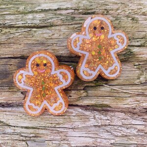 Könnte beinhalten: Zwei Lebkuchenmann-Anhänger mit weißem Zuckerguss und Glitzerdetails. Die Anhänger sind aus Harz gefertigt und haben eine glänzende Oberfläche.