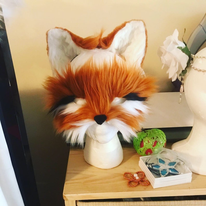 May include: A red and white fox fur mask with black eyes. The mask is on a white mannequin head.