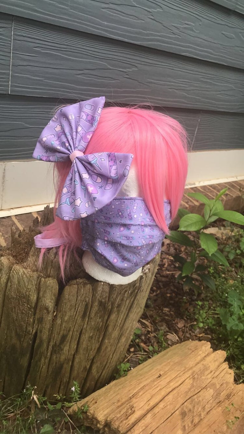 May include: A purple face mask with a pattern of syringes, bandages, and pills. The mask is worn by a mannequin with pink hair and a purple bow with the same pattern as the mask.