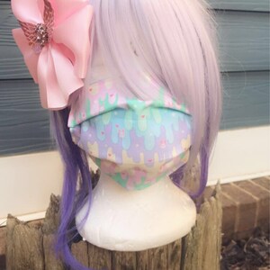 May include: A pastel pink and blue face mask with a pattern of melting ice cream cones and bunnies. The mask is on a white mannequin head with a pink bow and purple hair.