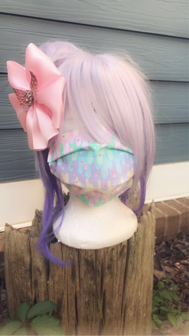 May include: A pastel pink and blue face mask with a pattern of melting ice cream cones and bunnies. The mask is on a white mannequin head with a pink bow and purple hair.