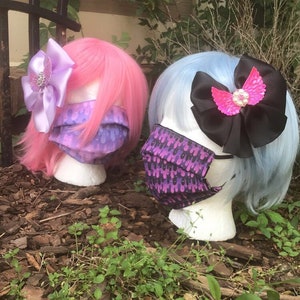 May include: Two mannequin heads with pink and blue wigs wearing purple and black patterned face masks. The mannequins are wearing decorative hair bows with rhinestones.
