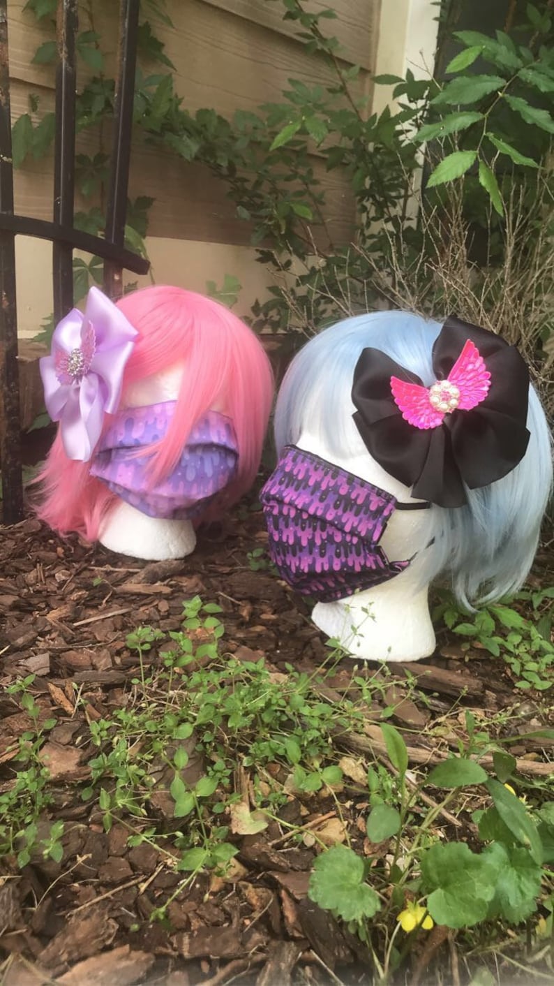 May include: Two mannequin heads with pink and blue wigs wearing purple and black patterned face masks. The mannequins are wearing decorative hair bows with rhinestones.