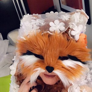 May include: A person wearing a white lace veil with a fox mask over their face. The mask is made of brown and white fur and has a black nose. The person is wearing a gold and brown dress with a floral pattern.