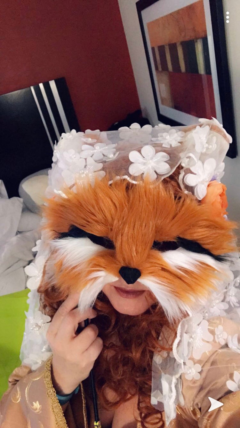 May include: A person wearing a white lace veil with a fox mask over their face. The mask is made of brown and white fur and has a black nose. The person is wearing a gold and brown dress with a floral pattern.