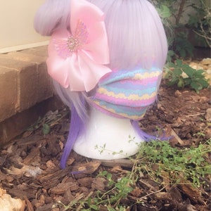 May include: A purple wig with a pink bow and a pastel-colored face mask with a wavy pattern. The wig and mask are on a mannequin head, which is sitting on a bed of brown wood chips.