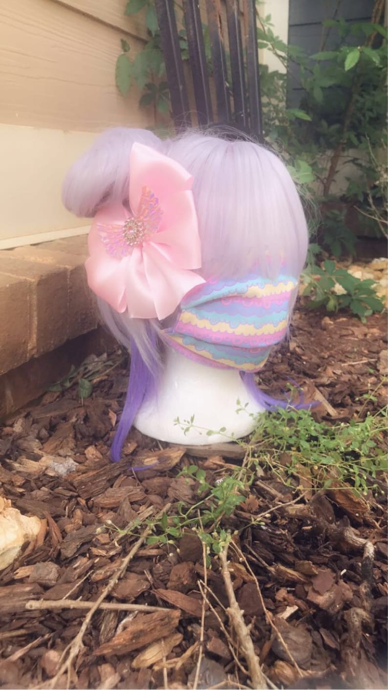 May include: A purple wig with a pink bow and a pastel-colored face mask with a wavy pattern. The wig and mask are on a mannequin head, which is sitting on a bed of brown wood chips.