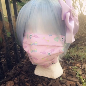 May include: A pink face mask with a pattern of bats, eyes, and bows. The mask is being worn by a mannequin with light blue hair and a purple bow.