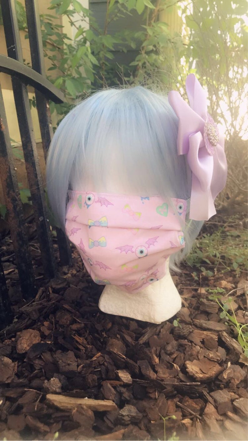 May include: A pink face mask with a pattern of bats, eyes, and bows. The mask is being worn by a mannequin with light blue hair and a purple bow.