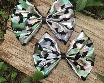May include: Two iridescent green and black bow hair clips with a bat pattern. The bows are on a wooden surface with green plants.