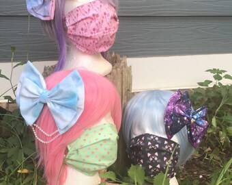 May include: Three mannequin heads with pink, purple, and blue wigs, each wearing a different patterned face mask and a large bow. The bows are in shades of blue and purple with a galaxy print. The mannequins are sitting on a wooden log in front of a green leafy background.