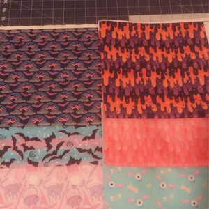 May include: Five different fabric squares with various Halloween-themed patterns. The top square has an orange and purple dripping pattern. The second square has a pink and white ombre pattern. The third square has a blue and pink pattern with bats. The fourth square has a pink and white pattern with witches' hats and cats. The bottom square has a blue and white pattern with candy corn and eyeballs.
