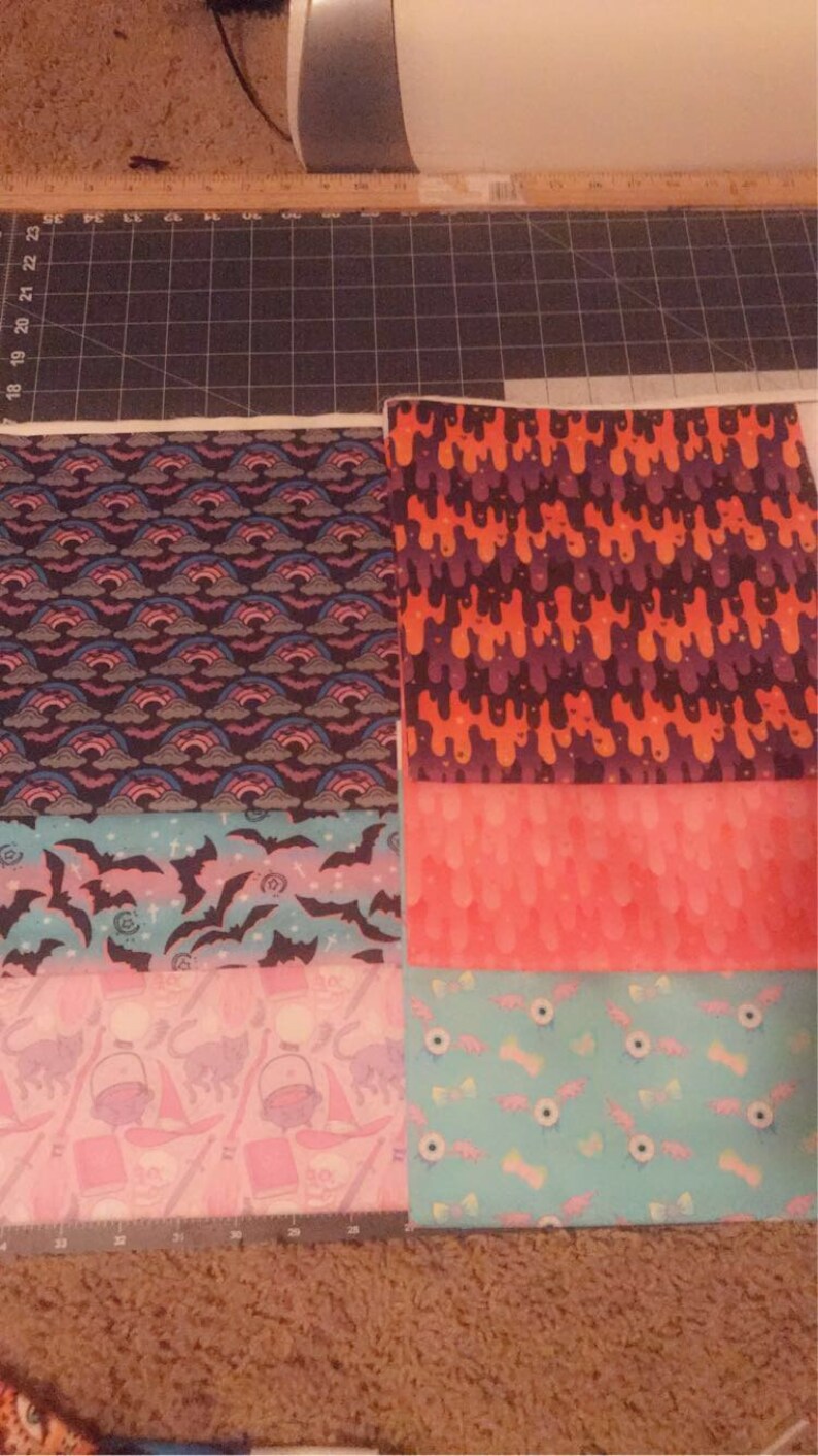 May include: Five different fabric squares with various Halloween-themed patterns. The top square has an orange and purple dripping pattern. The second square has a pink and white ombre pattern. The third square has a blue and pink pattern with bats. The fourth square has a pink and white pattern with witches' hats and cats. The bottom square has a blue and white pattern with candy corn and eyeballs.