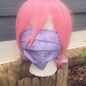 May include: A purple face mask with a white background and a pattern of syringes, pills, and bandages. The mask is on a mannequin head with pink hair.
