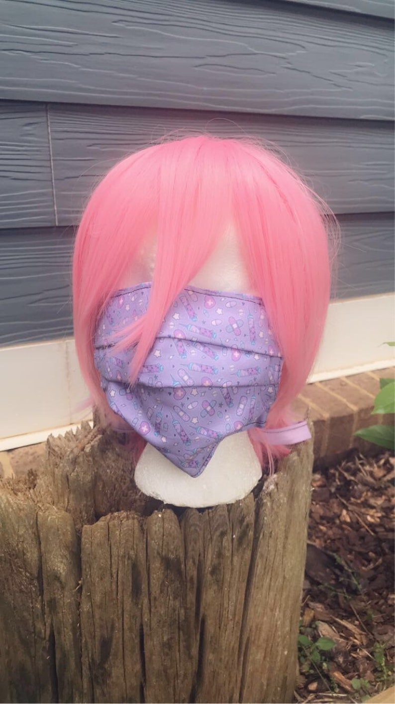 May include: A purple face mask with a white background and a pattern of syringes, pills, and bandages. The mask is on a mannequin head with pink hair.