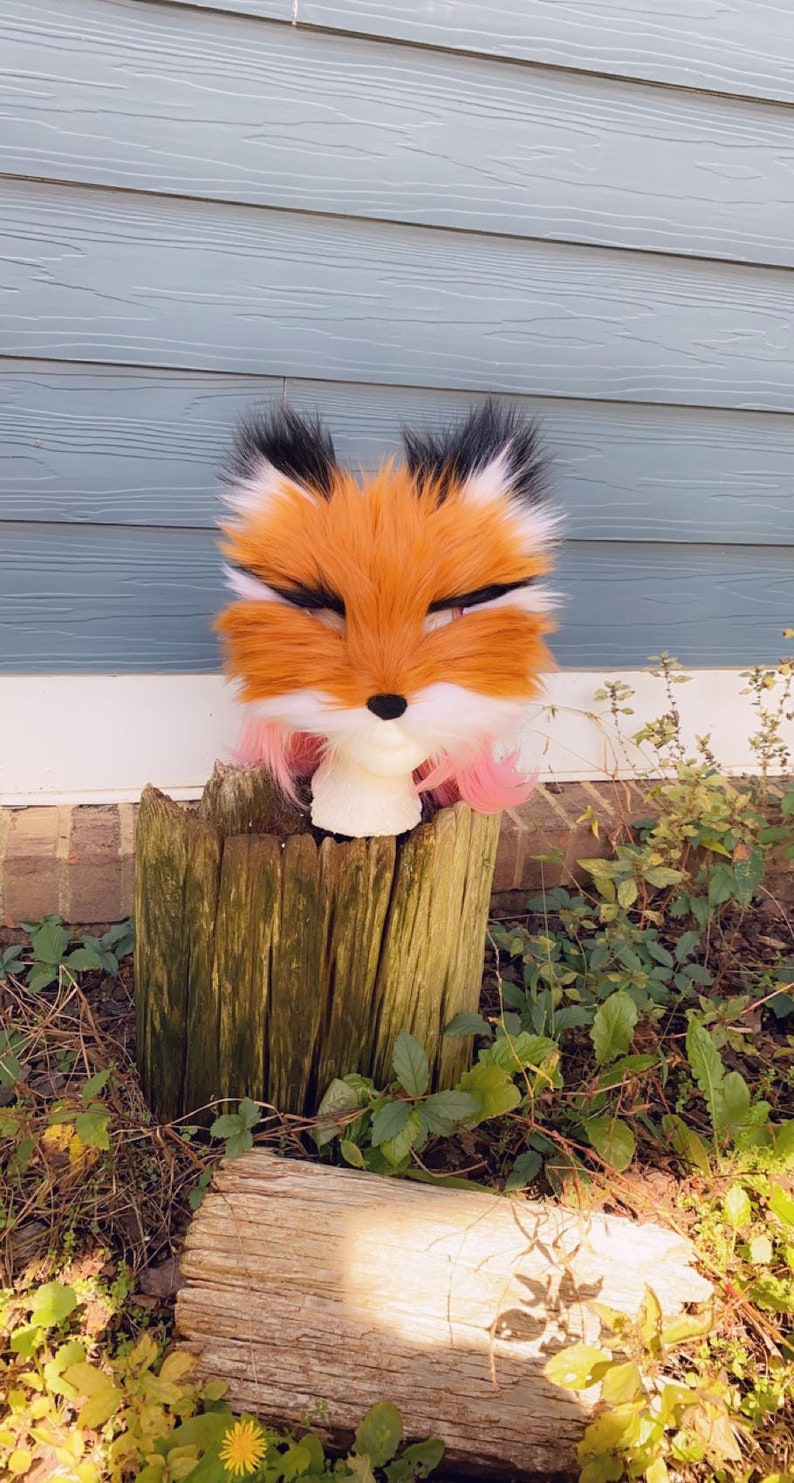 May include: A fox head fur costume with orange, white, and black fur. The costume is on a mannequin head with pink hair. The mannequin is sitting on a wooden stump.
