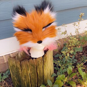 May include: A plush fox head with orange, white, and black fur. The head is sitting on a wooden stump with green moss and surrounded by green leaves.