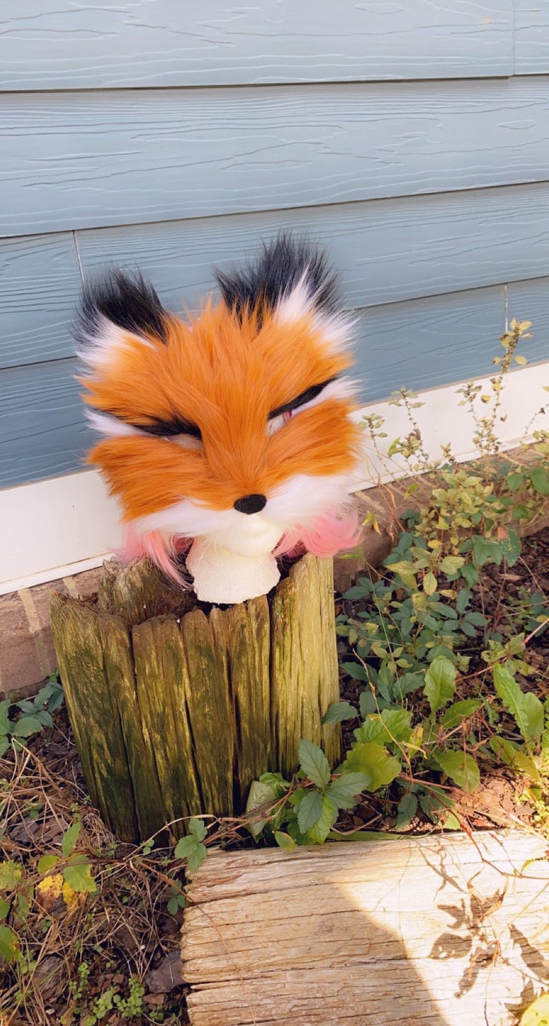 May include: A plush fox head with orange, white, and black fur. The head is sitting on a wooden stump with green moss and surrounded by green leaves.