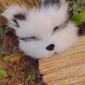 May include: A white and black furry animal mask with green eyes and a black nose. The mask is lying on a piece of weathered wood with green foliage in the background.