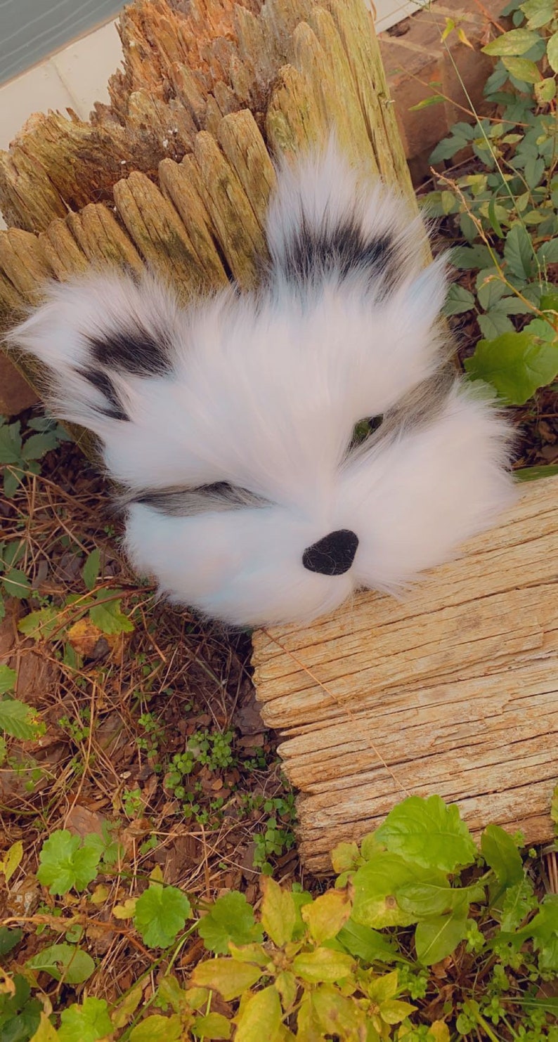 May include: A white and black furry animal mask with green eyes and a black nose. The mask is lying on a piece of weathered wood with green foliage in the background.
