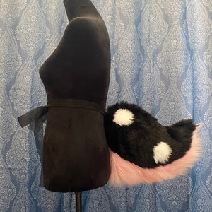 Peut inclure: Une queue en fausse fourrure noire et blanche à pois avec une pointe rose attachée à un mannequin noir.