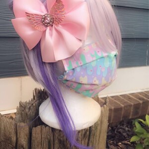 May include: A pink satin bow with a sparkly iridescent butterfly charm, worn on a purple wig with a pastel patterned face mask.
