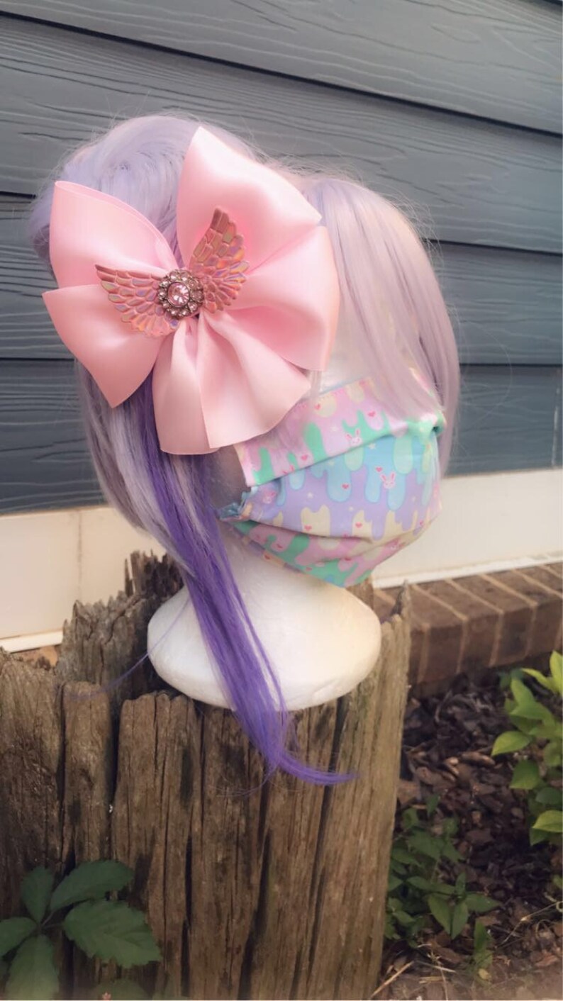 May include: A pink satin bow with a sparkly iridescent butterfly charm, worn on a purple wig with a pastel patterned face mask.