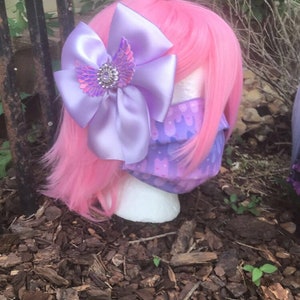 May include: A pink wig with a purple bow and a purple and pink patterned face mask. The bow has a sparkly iridescent butterfly design in the center.