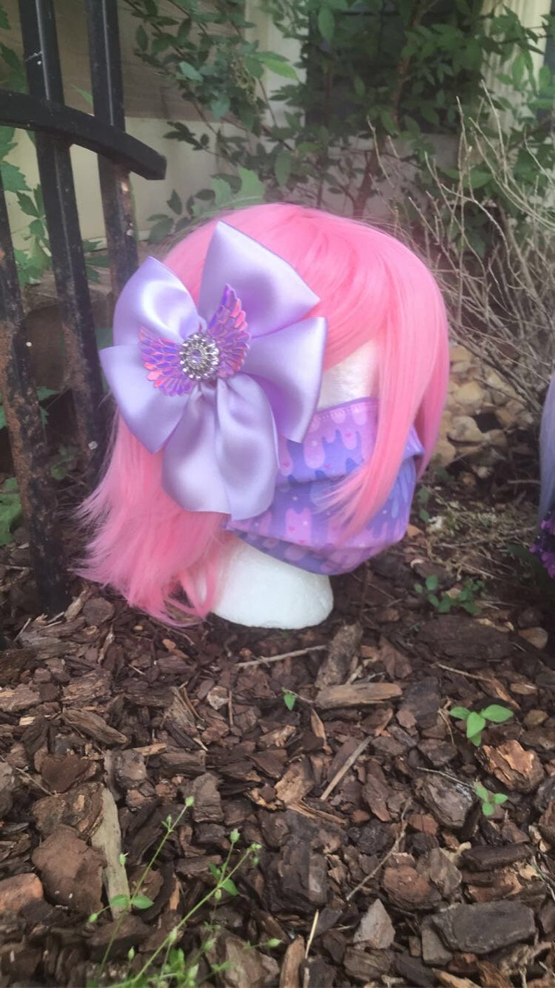 May include: A pink wig with a purple bow and a purple and pink patterned face mask. The bow has a sparkly iridescent butterfly design in the center.
