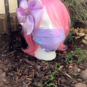 May include: A purple and pink face mask with a whimsical design featuring stars, hearts, and clouds. The mask is on a pink wig with a large purple bow.
