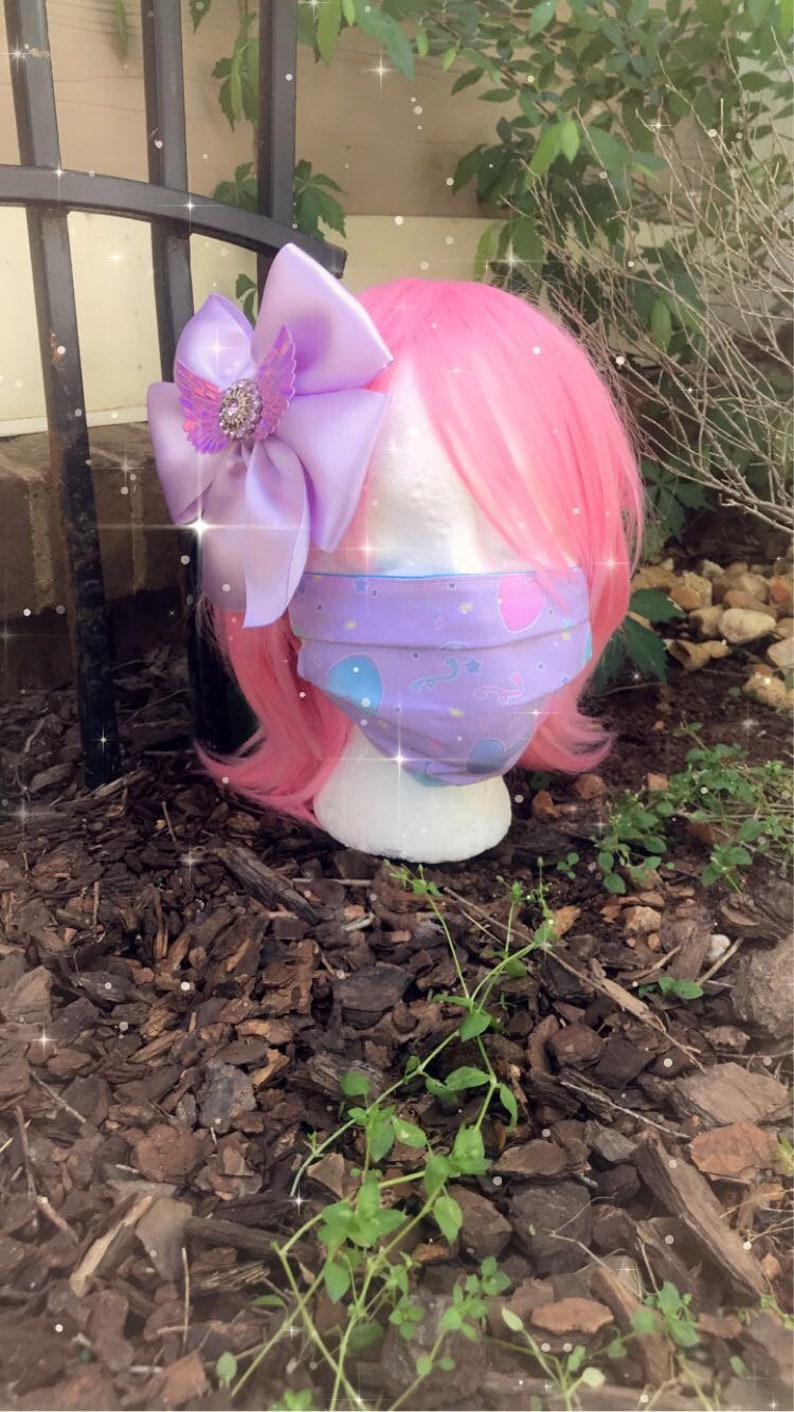 May include: A purple and pink face mask with a whimsical design featuring stars, hearts, and clouds. The mask is on a pink wig with a large purple bow.
