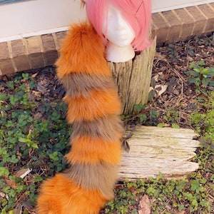 Op de afbeelding: Een rode panda staart kostuum accessoire met een pluche oranje en bruine gestreepte staart. De staart is gemaakt van imitatiebont en is bevestigd aan een hoofdband met rode panda oren.