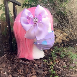 May include: A purple satin bow with a sparkly rhinestone and angel wings, attached to a pink wig with a purple and blue patterned face mask.
