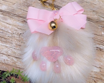 May include: A fluffy white faux fur keychain with a pink paw print design and a pink bow. The keychain has a gold bell charm.