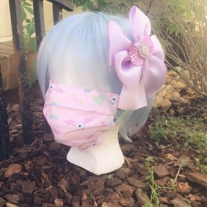 May include: A pink face mask with a pattern of eyes, bats, and hearts. The mask is on a mannequin with light blue hair and a lavender bow with a rhinestone embellishment.