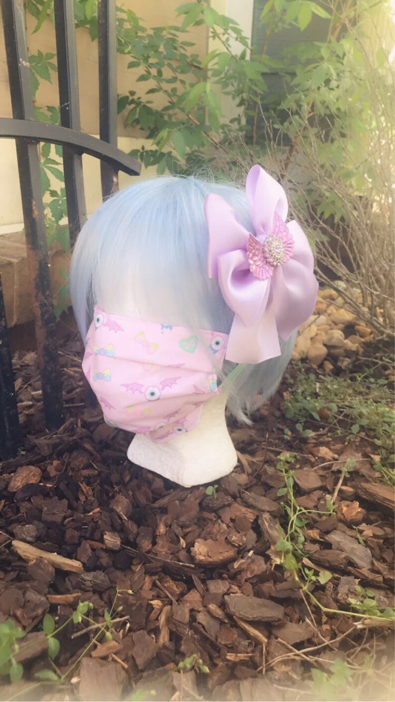 May include: A pink face mask with a pattern of eyes, bats, and hearts. The mask is on a mannequin with light blue hair and a lavender bow with a rhinestone embellishment.