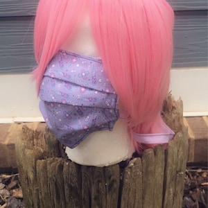 May include: A purple face mask with a pattern of syringes, bandages, and stars. The mask is on a mannequin head with pink hair.