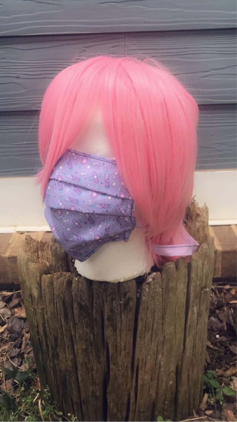 May include: A purple face mask with a pattern of syringes, bandages, and stars. The mask is on a mannequin head with pink hair.