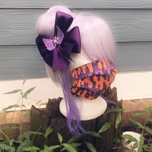 May include: A purple and orange patterned face mask with a purple bow with a sparkly butterfly accent in the center. The bow is attached to a purple wig.